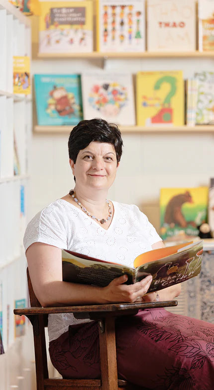 Photo of Kinder Books owner Anne Uebbing sitting in a chair with an open book.